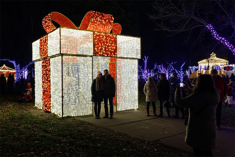 A large illuminated gift box display glowing with white and red lights at a nighttime holiday festival, with two people standing inside for a photo and others gathered around taking pictures among the surrounding light decorations.